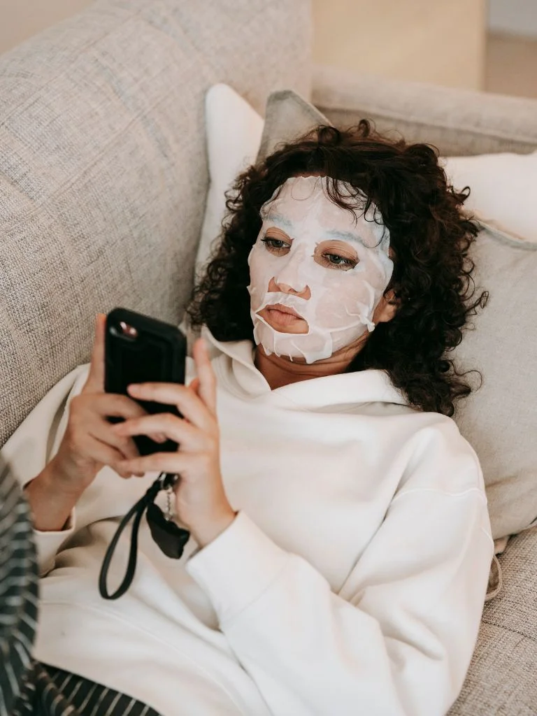 a woman scrolling on her phone with a sheet mask on her face
